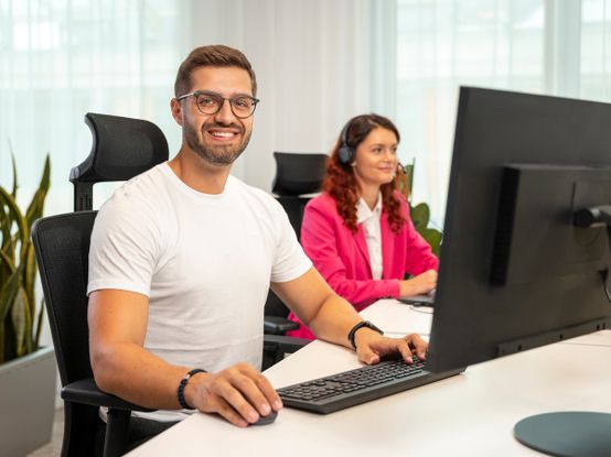 kolegyně se sluchátky a usmívající se kolega u monitoru