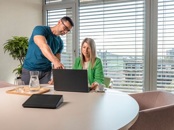 Muž a žena v jednací místnosti. Muž ukazuje ženě něco na obrazovce notebooku.