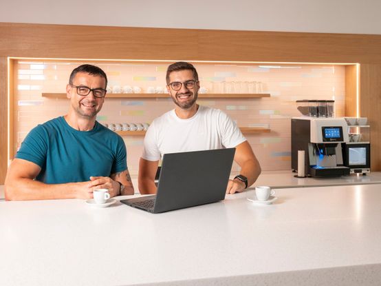 dva kolegové usmívající se u notebooku a kávy