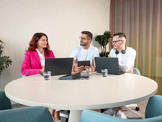 Dva muži a jedna žena sedí společně u stolu, usmívají se a baví se, zatímco pracují na notebooku.