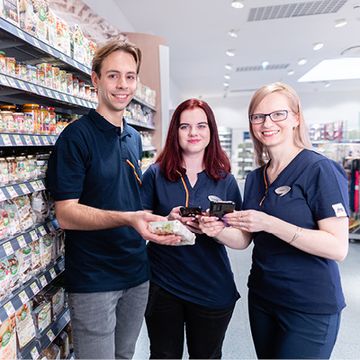 Tři spolupracovníci z prodejny dm, muž a dvě ženy, stojí u regálu, usmívají se a drží v ruce produkty dm.