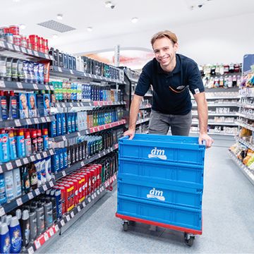 Prodavač projíždí v prodejně dm mezi regály s flatboxem plným zboží a usmívá se