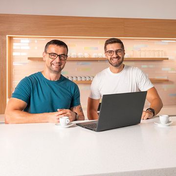 Dva spolupracovníci v kuchyňce stojí u notebooku a diskutují.