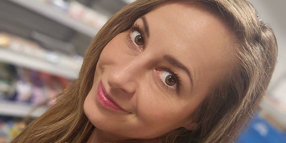 A woman with long brown hair smiles at the camera in a store setting, with shelves of products blurred in the background.