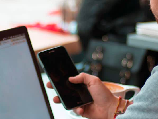 Un uomo tiene in mano uno smartphone mentre lavora al computer, con una tazza di caffè accanto.